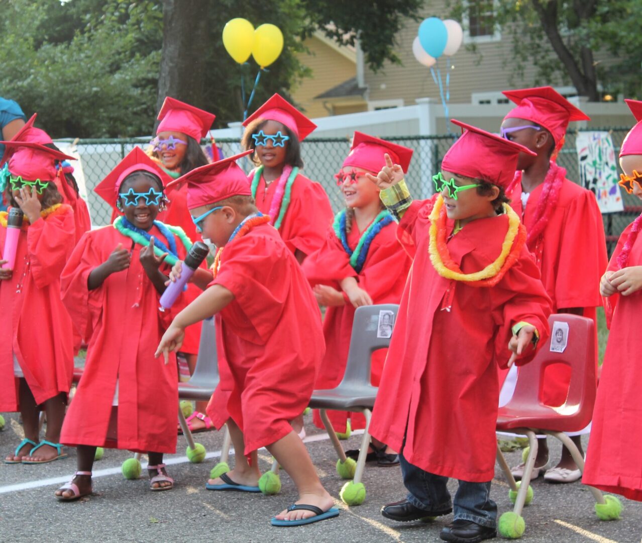 Children dressed in red graduation gowns and caps celebrate outdoors.