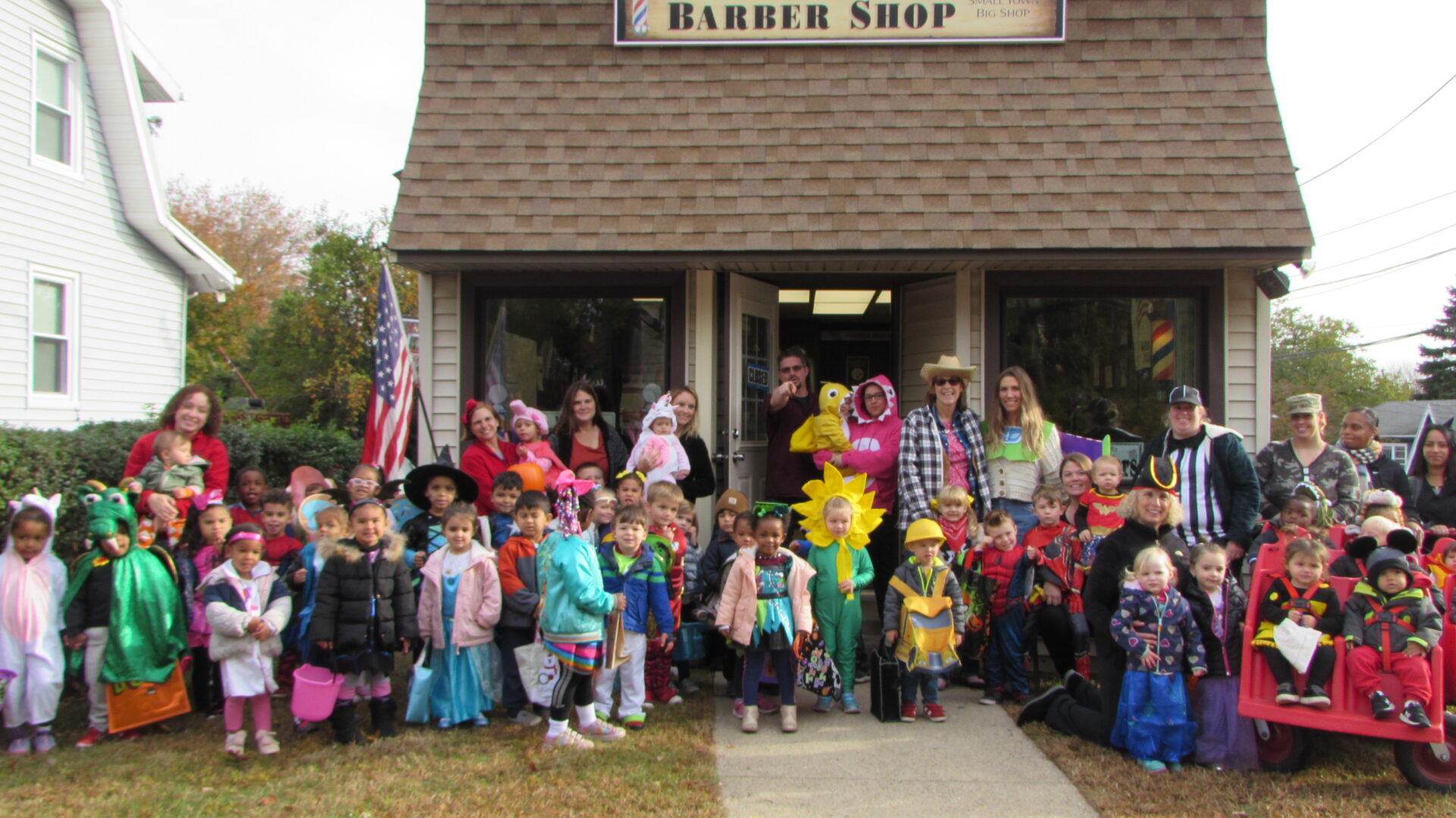 A large group of children and adults in colorful costumes outside a barber shop.