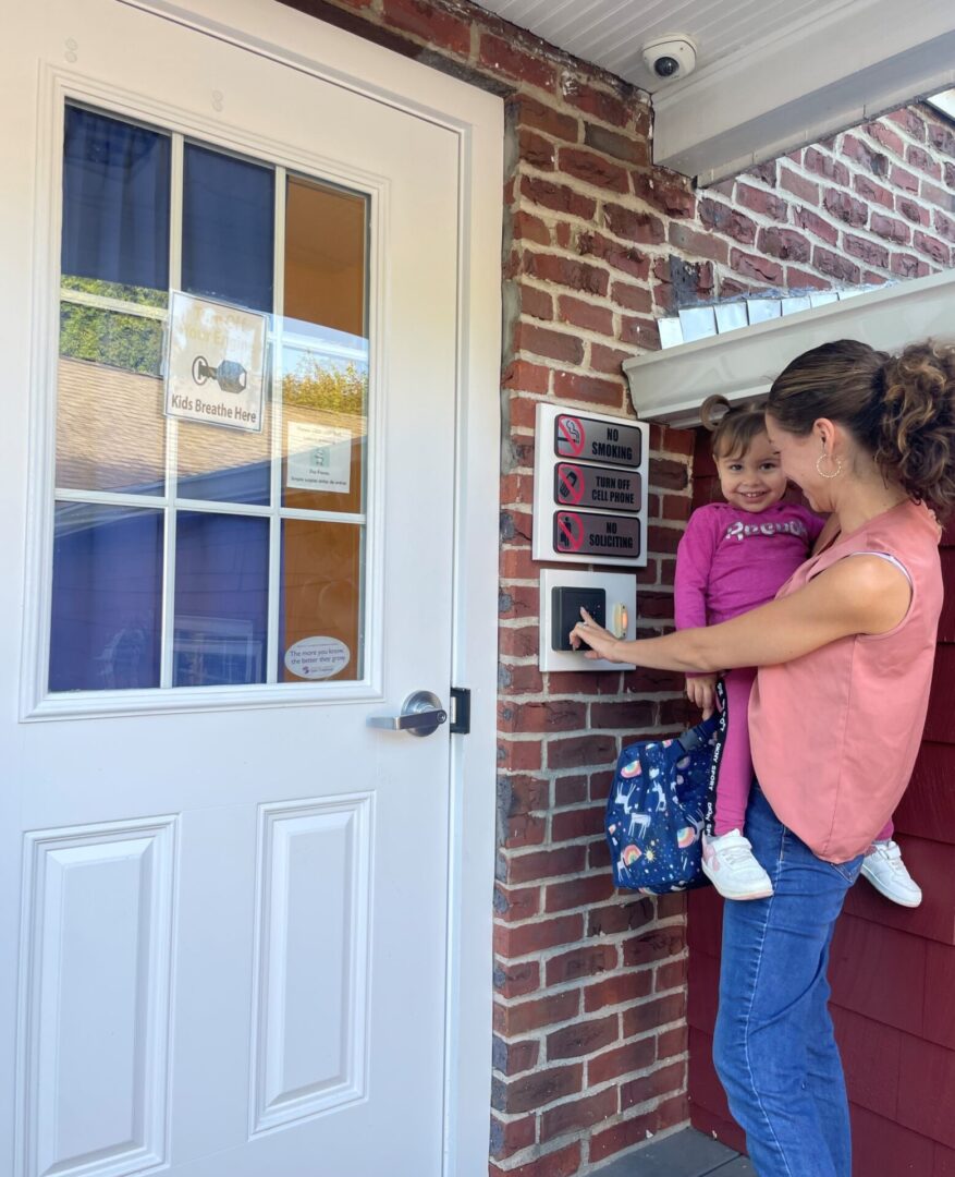 A woman holding a child opens a door with a keypad lock on a brick wall.